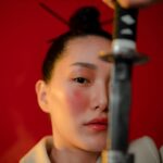 Close-up of a serious woman holding a katana in a studio shoot with a bold red background.